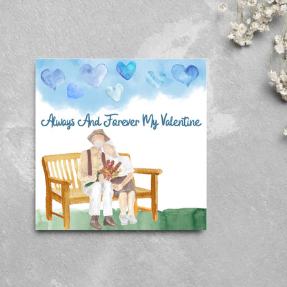 This beautiful Card has a watercolour style print with a couple sitting on a bench with flowers. Above them the blue sky has a series of cute watercolour hearts. This Cute card is perfect for valentines day for your wife or husband