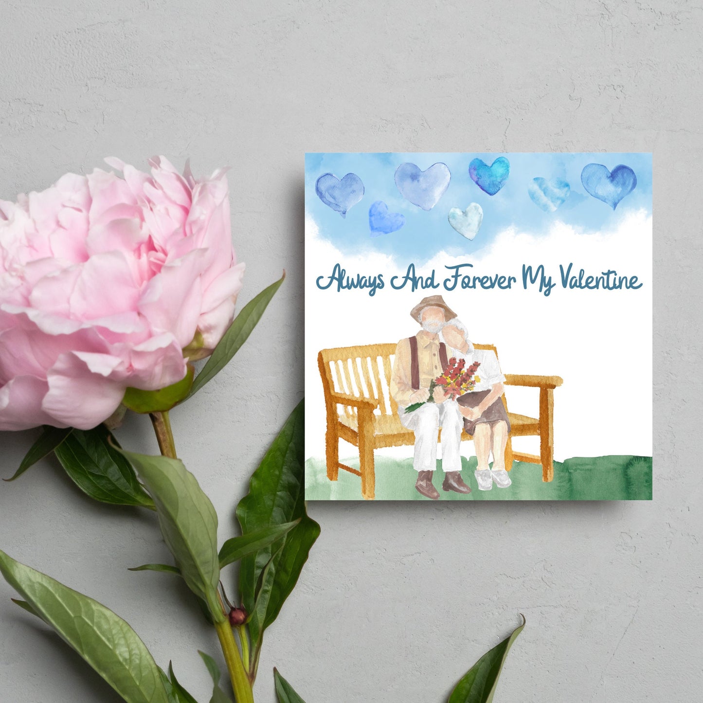 This beautiful Card has a watercolour style print with a couple sitting on a bench with flowers. Above them the blue sky has a series of cute watercolour hearts. This Cute card is perfect for valentines day for your wife or husband