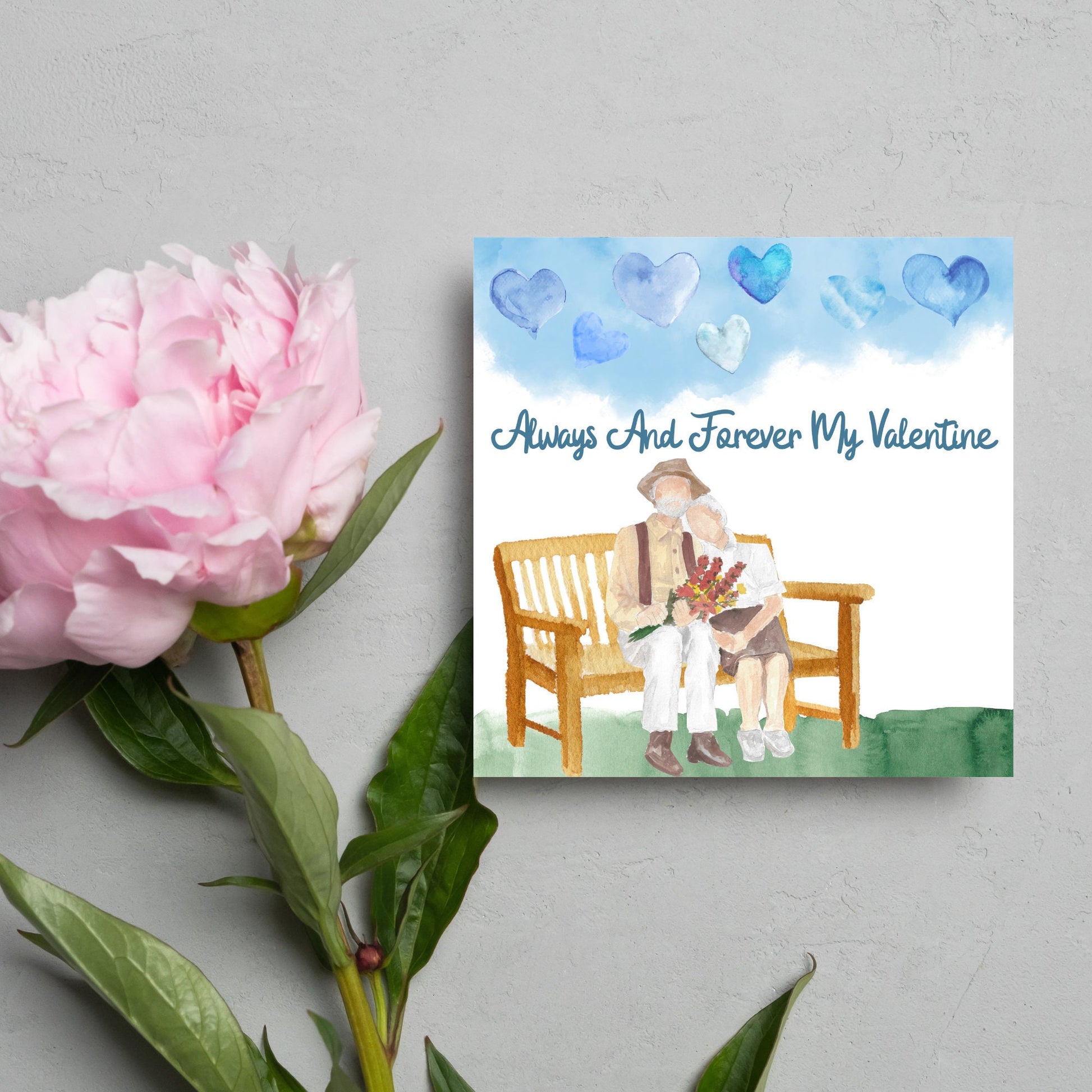 This beautiful Card has a watercolour style print with a couple sitting on a bench with flowers. Above them the blue sky has a series of cute watercolour hearts. This Cute card is perfect for valentines day for your wife or husband