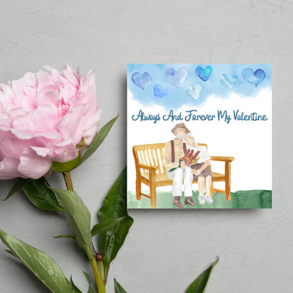 This beautiful Card has a watercolour style print with a couple sitting on a bench with flowers. Above them the blue sky has a series of cute watercolour hearts. This Cute card is perfect for valentines day for your wife or husband