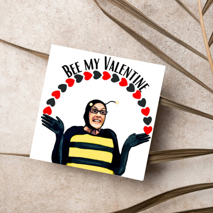 Square greetings card featuring Dean Pelton from the TV show Community, in one of his eccentric costumes dressed as a bee holding an arch of red and black hearts above his head with the text &#39;Bee my valentine&#39;
