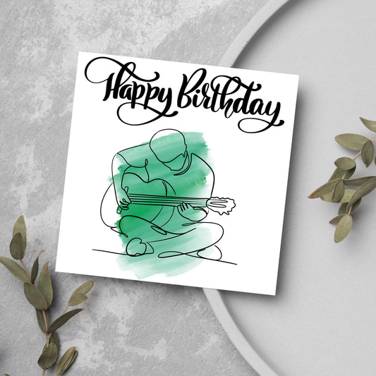 Square birthday card featuring a line drawing of a person sitting cross legged playing a guitar with a green watercolour style backdrop. The top of the card reads Happy Birthday in black swirly writing