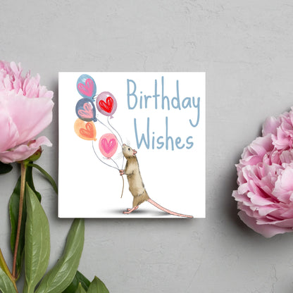 This square card features an illistration of a small mouse/rat holding some balloons with love hearts in a cute cartoon design. The card is captioned Birthday Wishes in a blue handwritten font. The design is on a white background