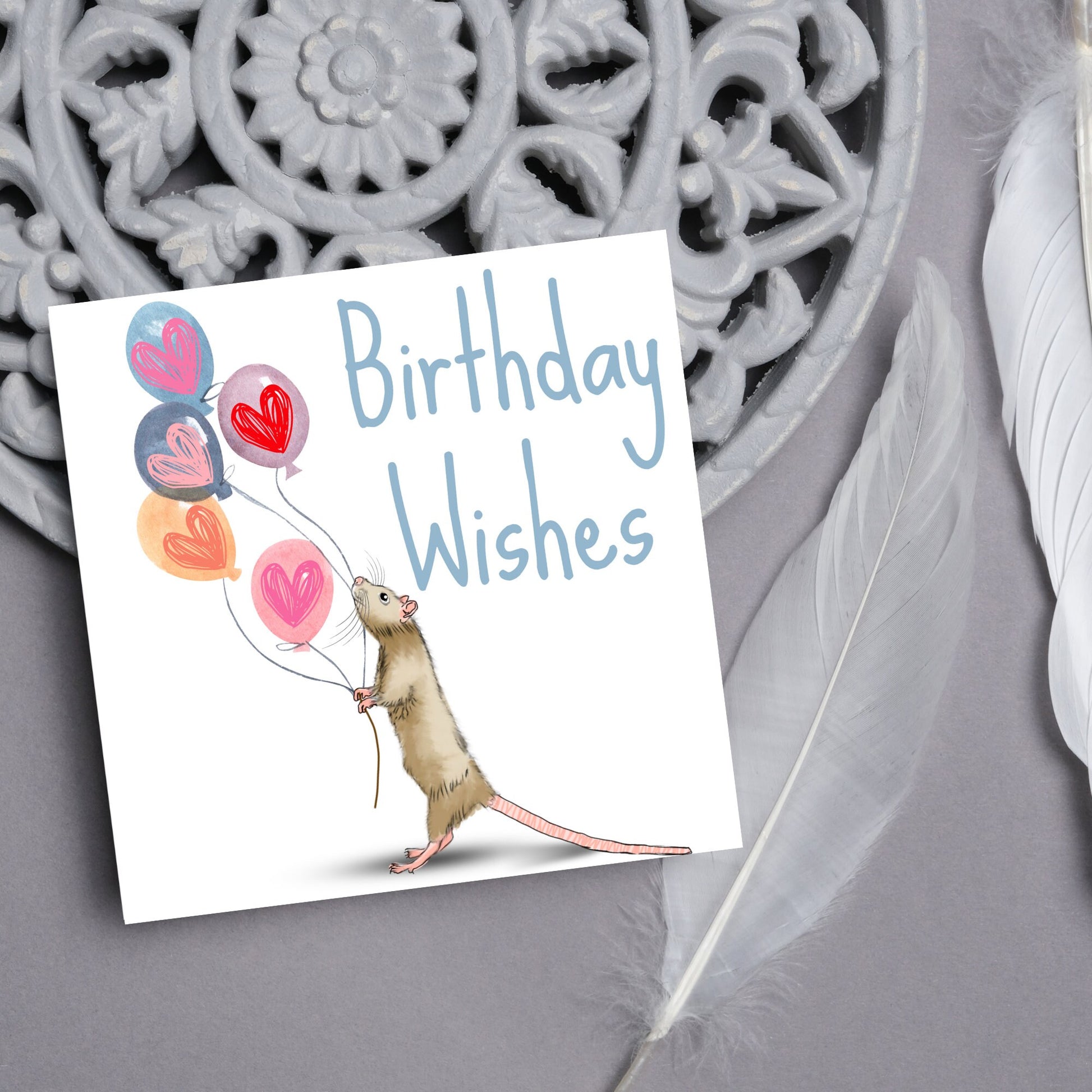 This square card features an illistration of a small mouse/rat holding some balloons with love hearts in a cute cartoon design. The card is captioned Birthday Wishes in a blue handwritten font. The design is on a white background