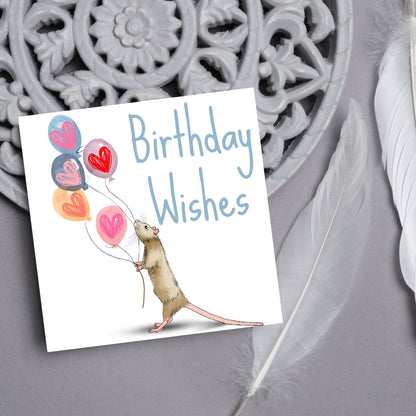 This square card features an illistration of a small mouse/rat holding some balloons with love hearts in a cute cartoon design. The card is captioned Birthday Wishes in a blue handwritten font. The design is on a white background