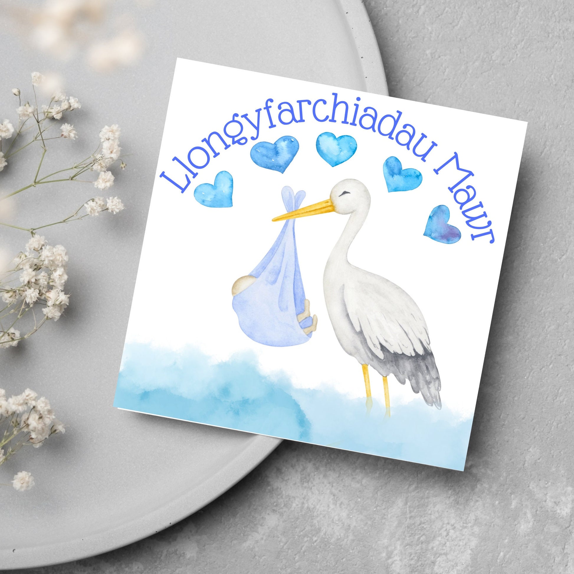 Image of a square greetings card with a stork delivering a baby in a blue blanket. THere are 5 blue watercolour hearts above the stork and the at the top is blue text &#39;llongyfarchiadau Mawr&#39; which translates from welsh as many congratulations