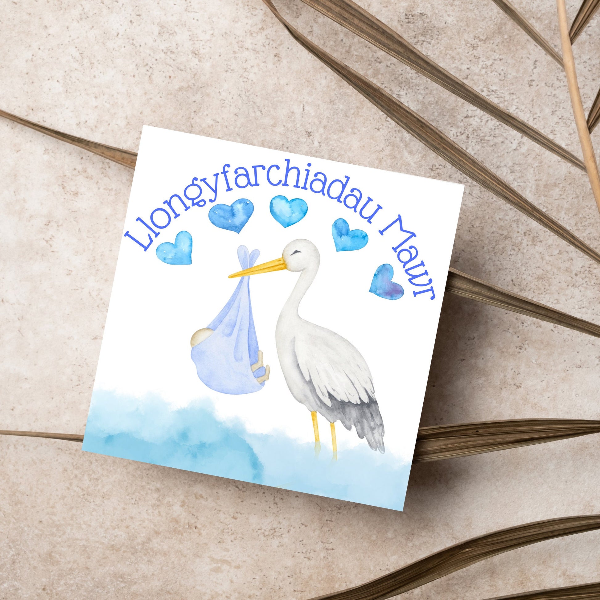 Image of a square greetings card with a stork delivering a baby in a blue blanket. THere are 5 blue watercolour hearts above the stork and the at the top is blue text &#39;llongyfarchiadau Mawr&#39; which translates from welsh as many congratulations
