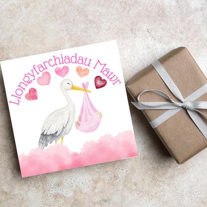 Image of a square greetings card with a stork delivering a baby in a pink blanket. There are 5 pink watercolour hearts above the stork and the at the top is pink text &#39;llongyfarchiadau Mawr&#39; which translates from welsh as many congratulations