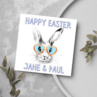 Square card with a simple watercolour style illustration of a white rabbit wearing orange and blue heart shaped sunglasses. The text reads &#39;Happy Easter Jane and Paul&#39;