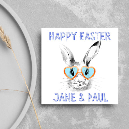 Square card with a simple watercolour style illustration of a white rabbit wearing orange and blue heart shaped sunglasses. The text reads &#39;Happy Easter Jane and Paul&#39;