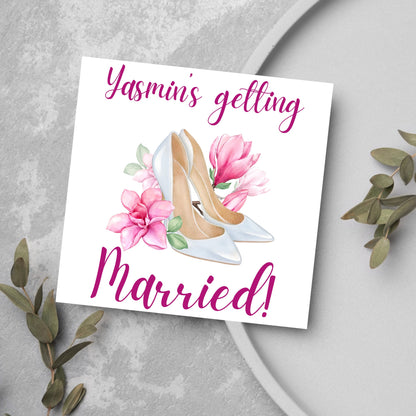Square card with a watercolour image of white wedding shoes surrounded by pink flowers with pink text that reads Yasmin&#39;s getting married in calligraphy style font