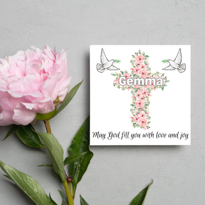 Square white card with a cross made of pink roses at the centre with the chosen name written across in white. either side of the cross is a white dove flying. At the bottom the text &#39;may god fill you with love and joy&#39; is written