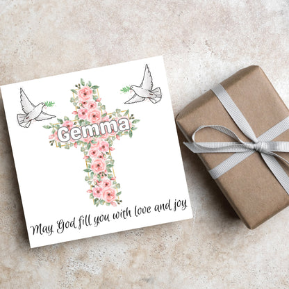 Square white card with a cross made of pink roses at the centre with the chosen name written across in white. either side of the cross is a white dove flying. At the bottom the text &#39;may god fill you with love and joy&#39; is written