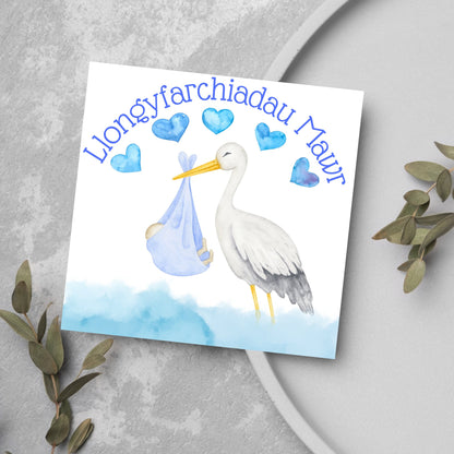 Image of a square greetings card with a stork delivering a baby in a blue blanket. THere are 5 blue watercolour hearts above the stork and the at the top is blue text &#39;llongyfarchiadau Mawr&#39; which translates from welsh as many congratulations