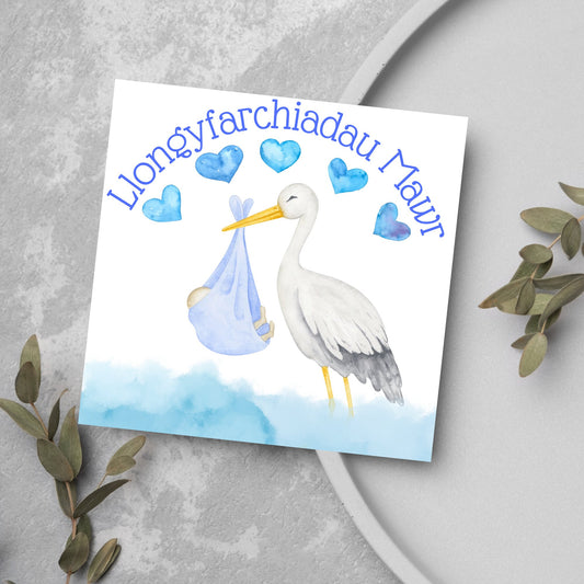 Image of a square greetings card with a stork delivering a baby in a blue blanket. THere are 5 blue watercolour hearts above the stork and the at the top is blue text &#39;llongyfarchiadau Mawr&#39; which translates from welsh as many congratulations