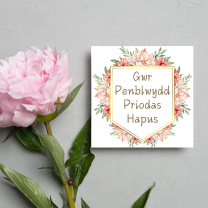 Image of a square wedding anniversary card with &#39;gwr penblwydd priodas hapus&#39; in a subtle handwritten style font, which translates to &#39;happy wedding anniversary&#39; in welsh. the text is against a white banner surrounded by pink flowers