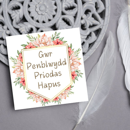 Image of a square wedding anniversary card with &#39;gwr penblwydd priodas hapus&#39; in a subtle handwritten style font, which translates to &#39;happy wedding anniversary&#39; in welsh. the text is against a white banner surrounded by pink flowers