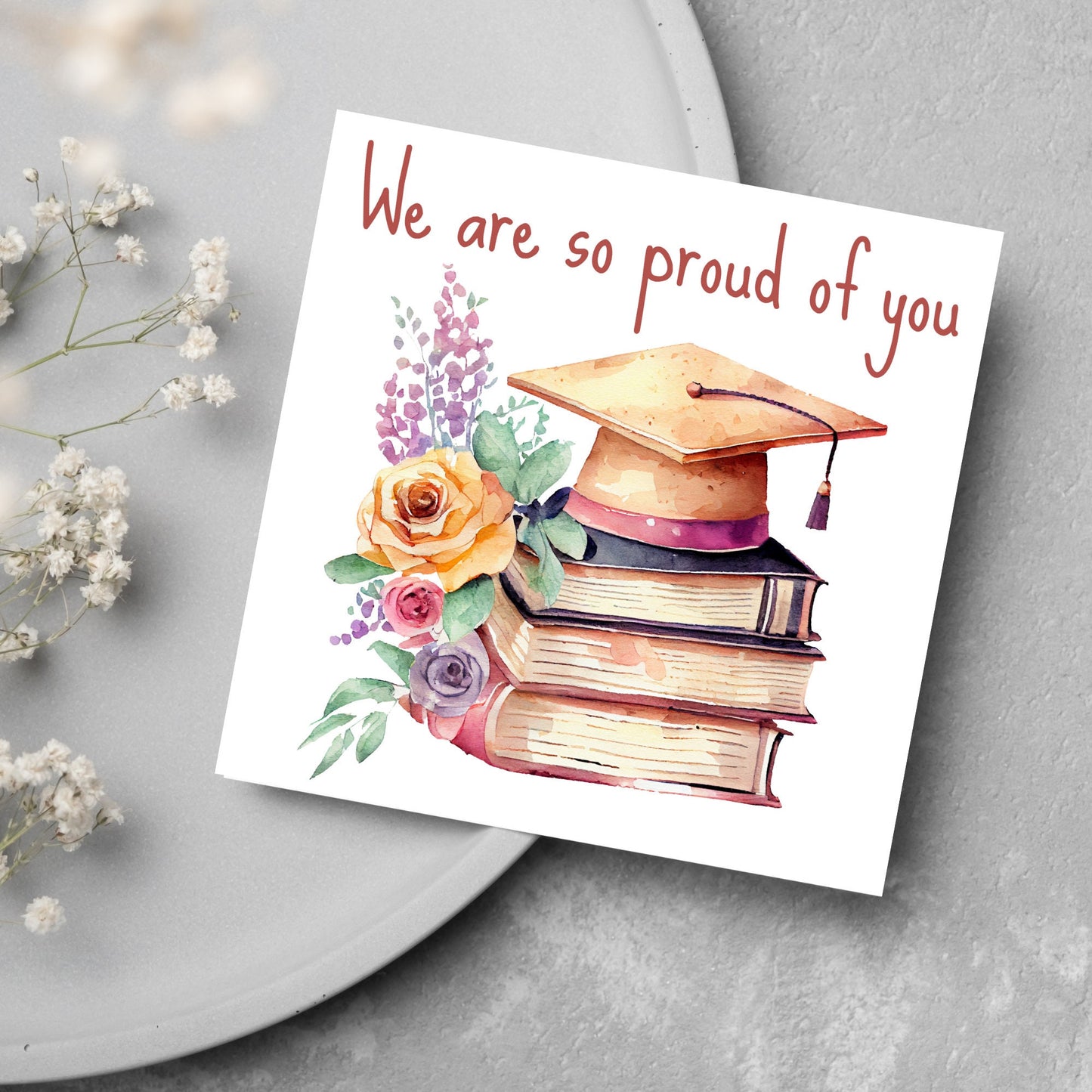Image of a square graduation greeting card featuring a watercolour style image of a graduation cap on top of a stack of 3 books next to a floral arrangement. Above the image is the text &#39;we are so proud of you&#39; in a handwritten font