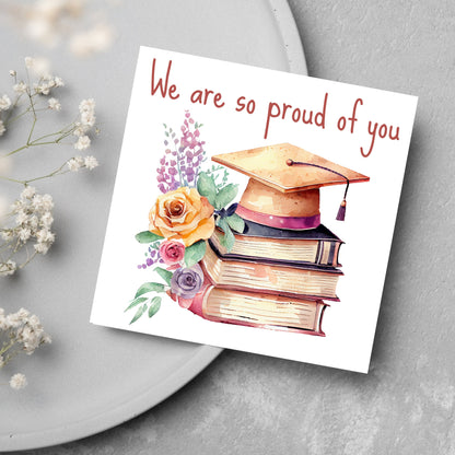 Image of a square graduation greeting card featuring a watercolour style image of a graduation cap on top of a stack of 3 books next to a floral arrangement. Above the image is the text &#39;we are so proud of you&#39; in a handwritten font