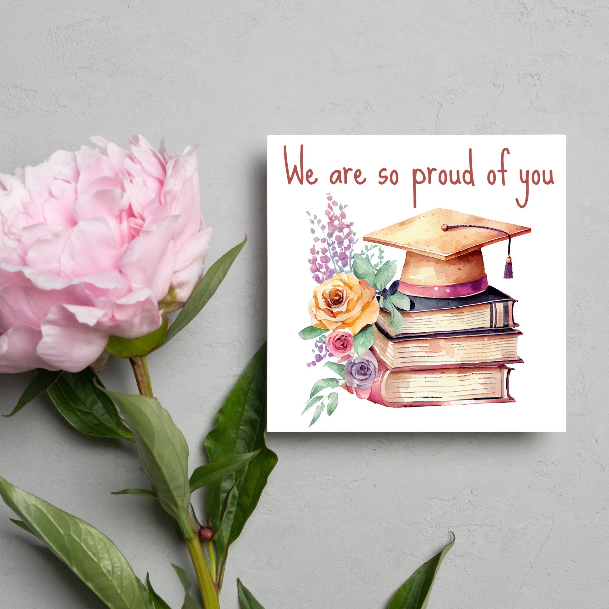 Image of a square graduation greeting card featuring a watercolour style image of a graduation cap on top of a stack of 3 books next to a floral arrangement. Above the image is the text &#39;we are so proud of you&#39; in a handwritten font