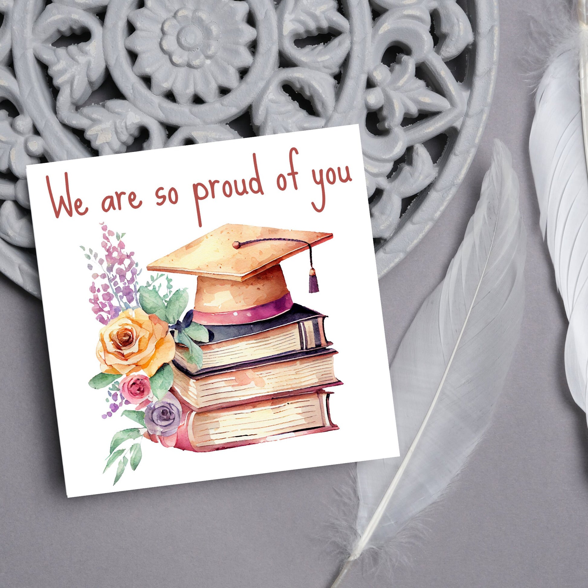 Image of a square graduation greeting card featuring a watercolour style image of a graduation cap on top of a stack of 3 books next to a floral arrangement. Above the image is the text &#39;we are so proud of you&#39; in a handwritten font