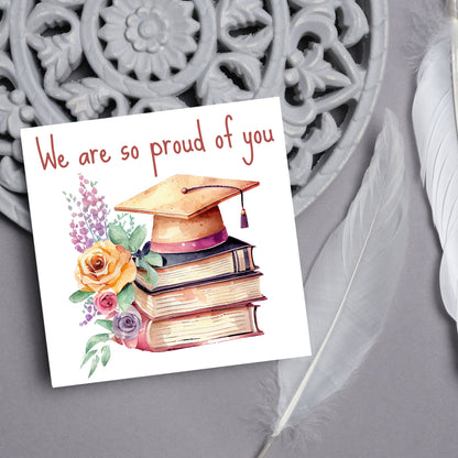 Image of a square graduation greeting card featuring a watercolour style image of a graduation cap on top of a stack of 3 books next to a floral arrangement. Above the image is the text &#39;we are so proud of you&#39; in a handwritten font