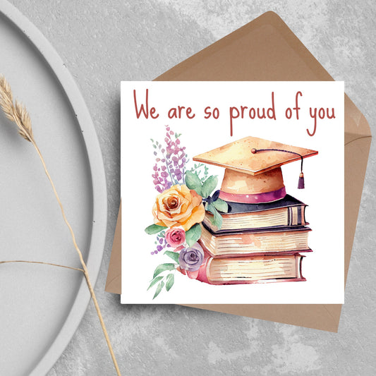 Image of a square graduation greeting card featuring a watercolour style image of a graduation cap on top of a stack of 3 books next to a floral arrangement. Above the image is the text &#39;we are so proud of you&#39; in a handwritten font