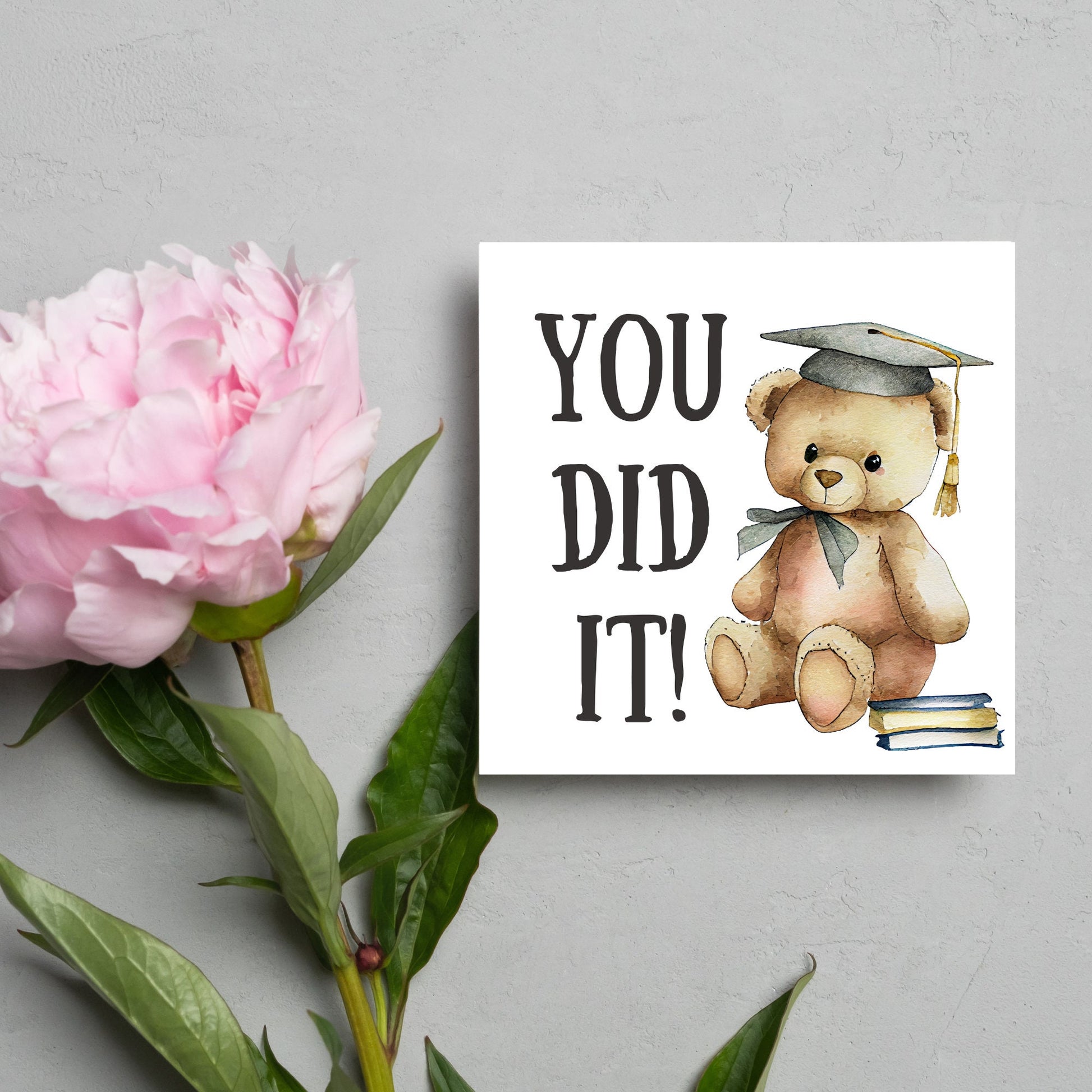 image of a square graduation greetings card featuring a watercolour illustration of a teddy bear wearing a graduation cap next to a pile of books. on the left of the bear is large black handwritten style text that reads &#39;you did it!&#39;