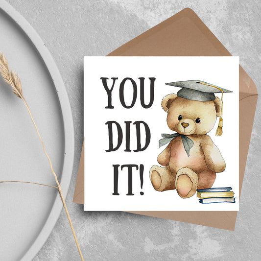 image of a square graduation greetings card featuring a watercolour illustration of a teddy bear wearing a graduation cap next to a pile of books. on the left of the bear is large black handwritten style text that reads &#39;you did it!&#39;