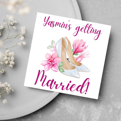 Square card with a watercolour image of white wedding shoes surrounded by pink flowers with pink text that reads Yasmin&#39;s getting married in calligraphy style font