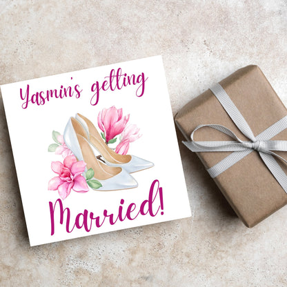 Square card with a watercolour image of white wedding shoes surrounded by pink flowers with pink text that reads Yasmin&#39;s getting married in calligraphy style font