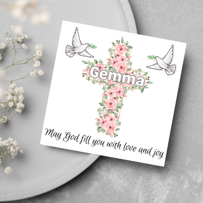 Square white card with a cross made of pink roses at the centre with the chosen name written across in white. either side of the cross is a white dove flying. At the bottom the text &#39;may god fill you with love and joy&#39; is written