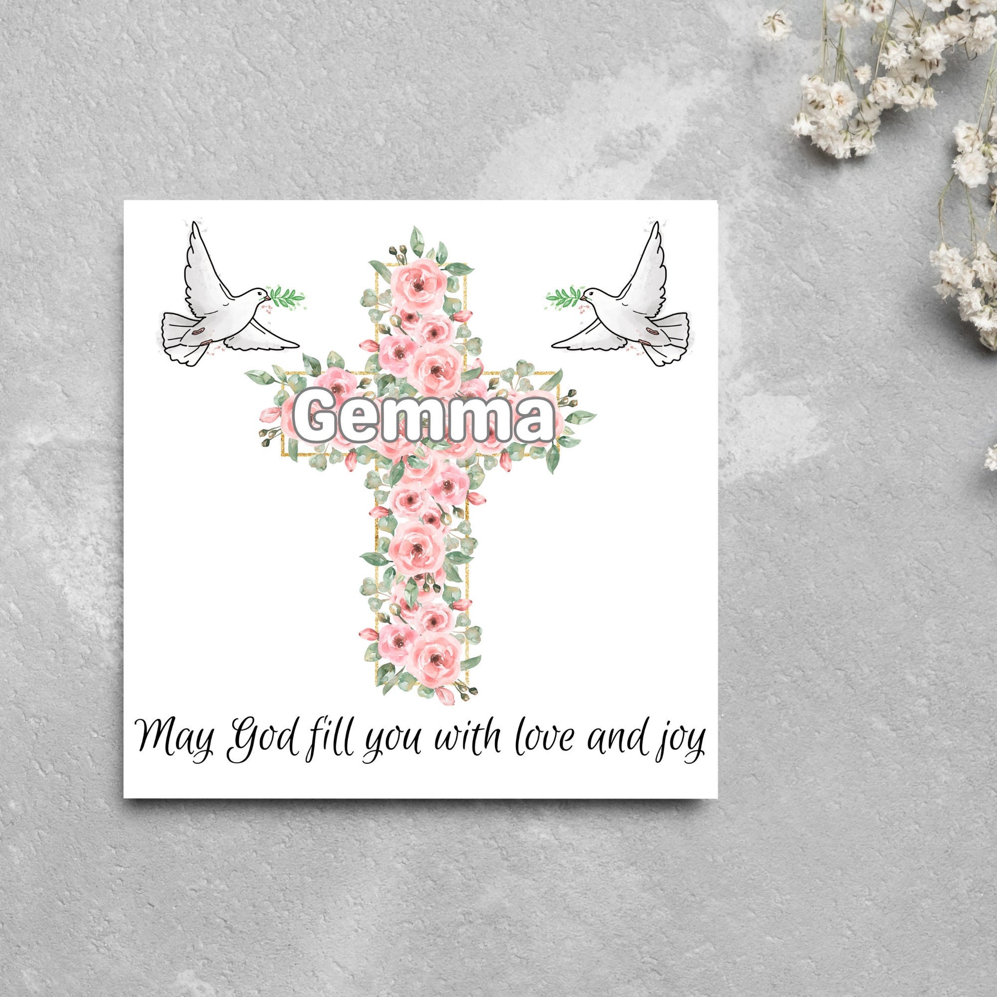 Square white card with a cross made of pink roses at the centre with the chosen name written across in white. either side of the cross is a white dove flying. At the bottom the text &#39;may god fill you with love and joy&#39; is written