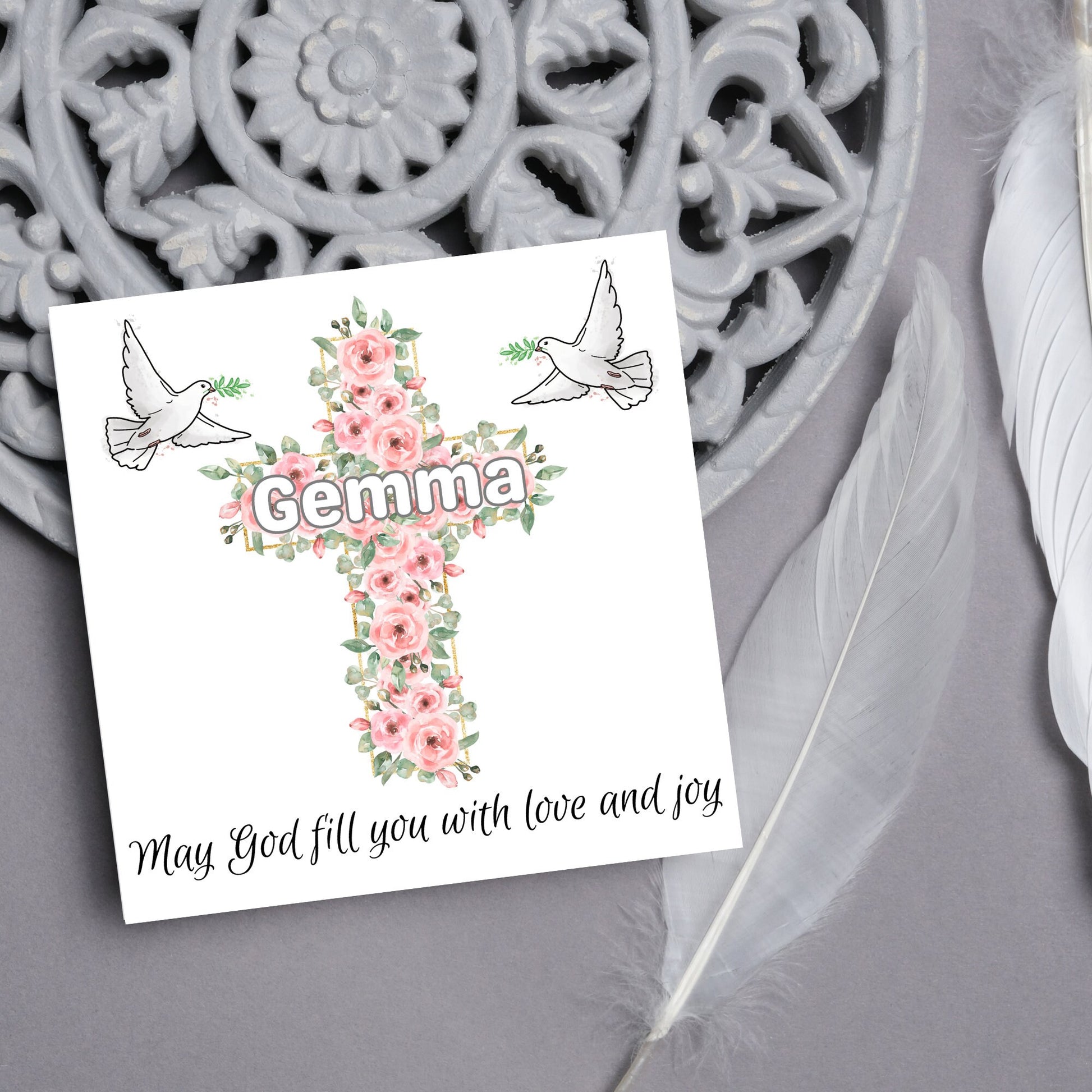 Square white card with a cross made of pink roses at the centre with the chosen name written across in white. either side of the cross is a white dove flying. At the bottom the text &#39;may god fill you with love and joy&#39; is written