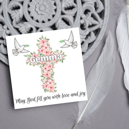 Square white card with a cross made of pink roses at the centre with the chosen name written across in white. either side of the cross is a white dove flying. At the bottom the text &#39;may god fill you with love and joy&#39; is written