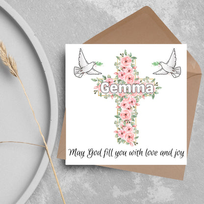 Square white card with a cross made of pink roses at the centre with the chosen name written across in white. either side of the cross is a white dove flying. At the bottom the text &#39;may god fill you with love and joy&#39; is written