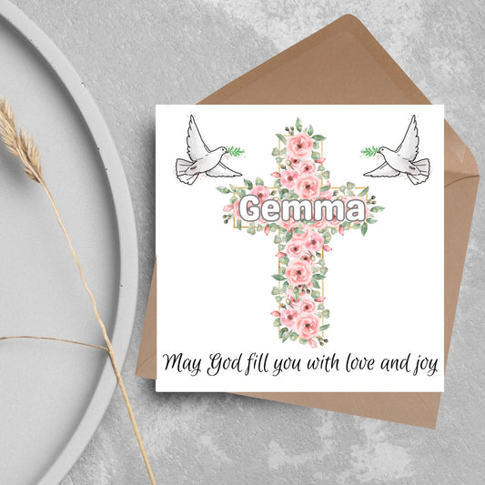 Square white card with a cross made of pink roses at the centre with the chosen name written across in white. either side of the cross is a white dove flying. At the bottom the text &#39;may god fill you with love and joy&#39; is written