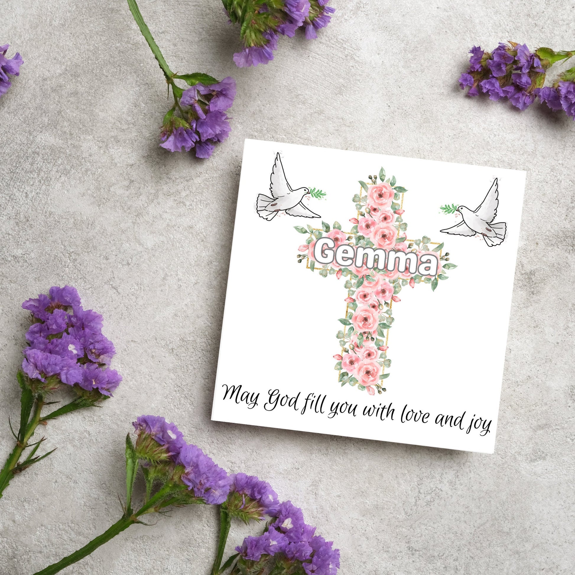 Square white card with a cross made of pink roses at the centre with the chosen name written across in white. either side of the cross is a white dove flying. At the bottom the text &#39;may god fill you with love and joy&#39; is written