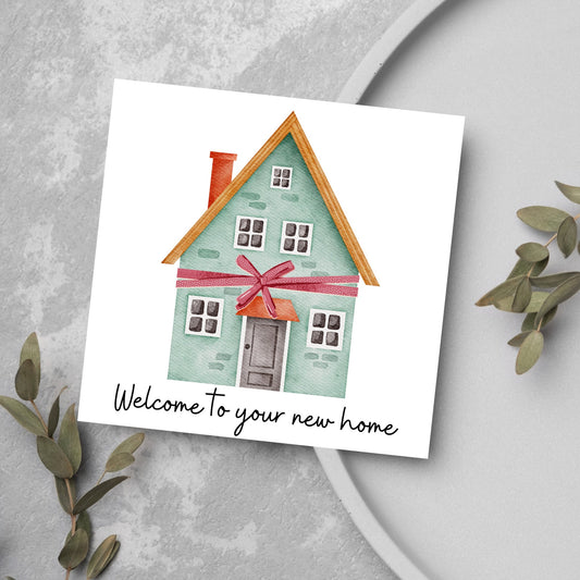 Square card with white background and a watercolour style illlustration of a green three storey house with a red bow wrapped around it. Underneath the house ina script font are the words &#39;welcome to your new home&#39;