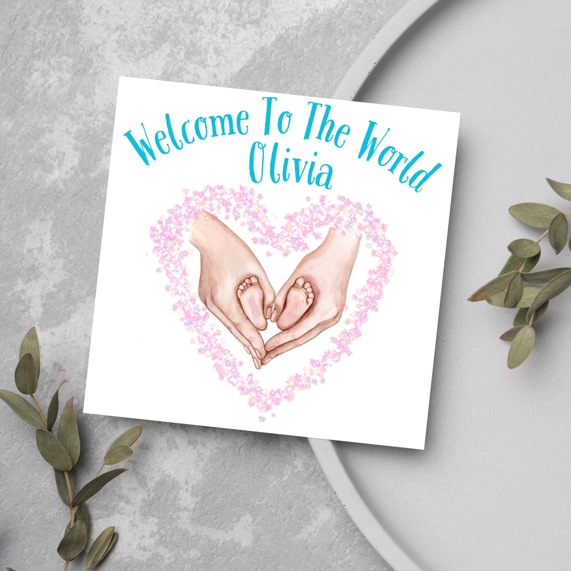 image of a square card with an illustration of hands holding newborn feet in a heart shape surrounded by pink flowers. Above the image is blue text that reads &#39;welcome to the world olivia&#39; in a handwritten style font
