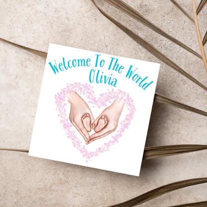 image of a square card with an illustration of hands holding newborn feet in a heart shape surrounded by pink flowers. Above the image is blue text that reads &#39;welcome to the world olivia&#39; in a handwritten style font