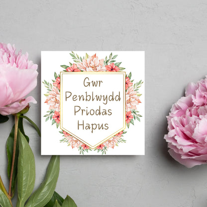 Image of a square wedding anniversary card with &#39;gwr penblwydd priodas hapus&#39; in a subtle handwritten style font, which translates to &#39;happy wedding anniversary&#39; in welsh. the text is against a white banner surrounded by pink flowers