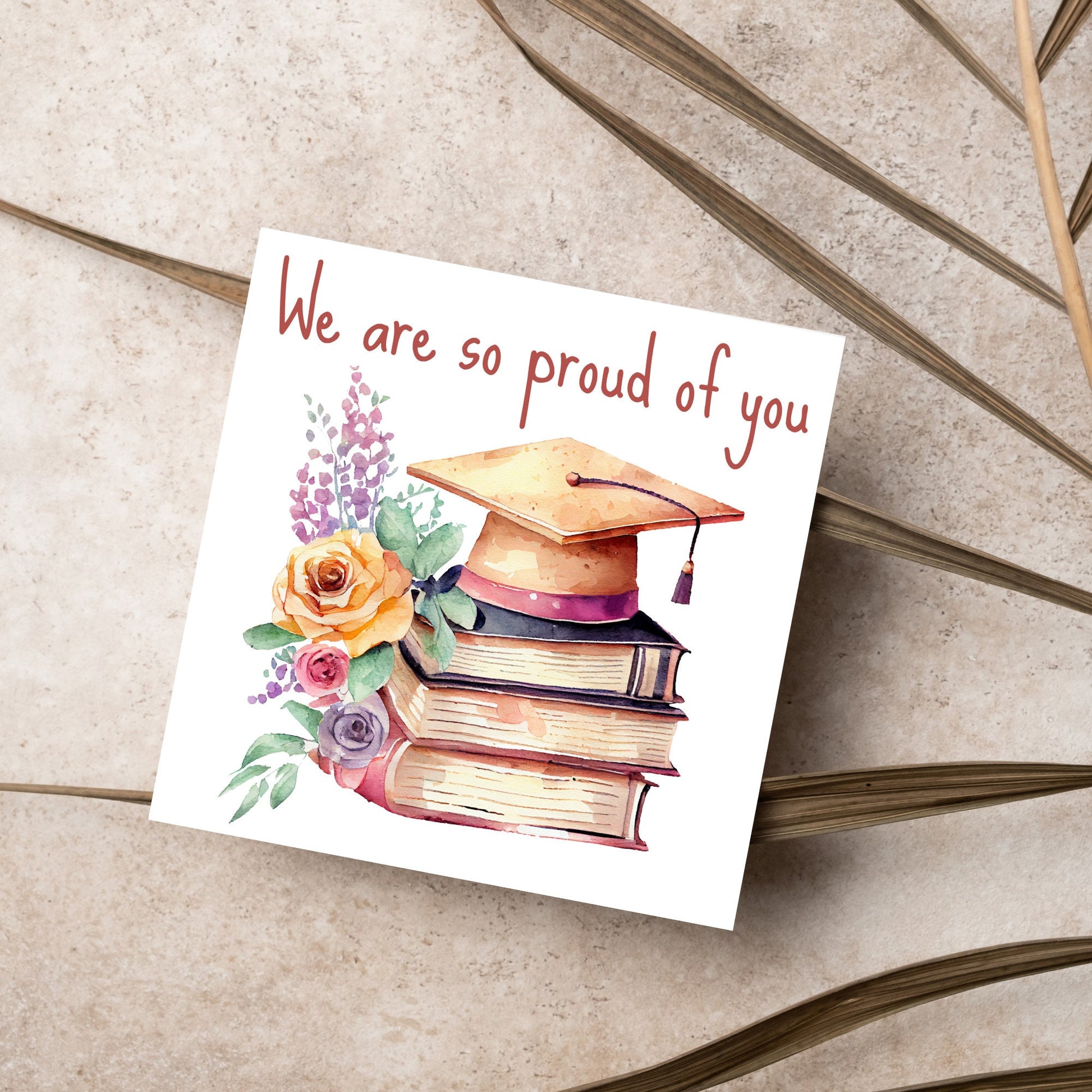 Image of a square graduation greeting card featuring a watercolour style image of a graduation cap on top of a stack of 3 books next to a floral arrangement. Above the image is the text &#39;we are so proud of you&#39; in a handwritten font