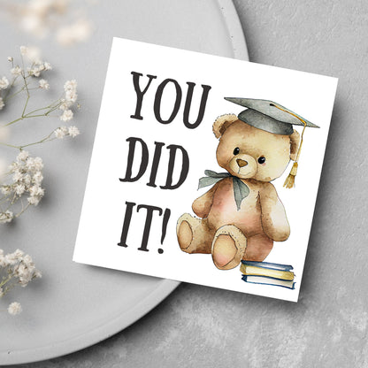 image of a square graduation greetings card featuring a watercolour illustration of a teddy bear wearing a graduation cap next to a pile of books. on the left of the bear is large black handwritten style text that reads &#39;you did it!&#39;