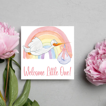 square new baby card with a flying cartoon stork delivering a swaddled baby against a watercolour rainbow in pastel shades. A cursive style text reads &#39;welcome little one&#39; in a light pink font at the bottom of the card