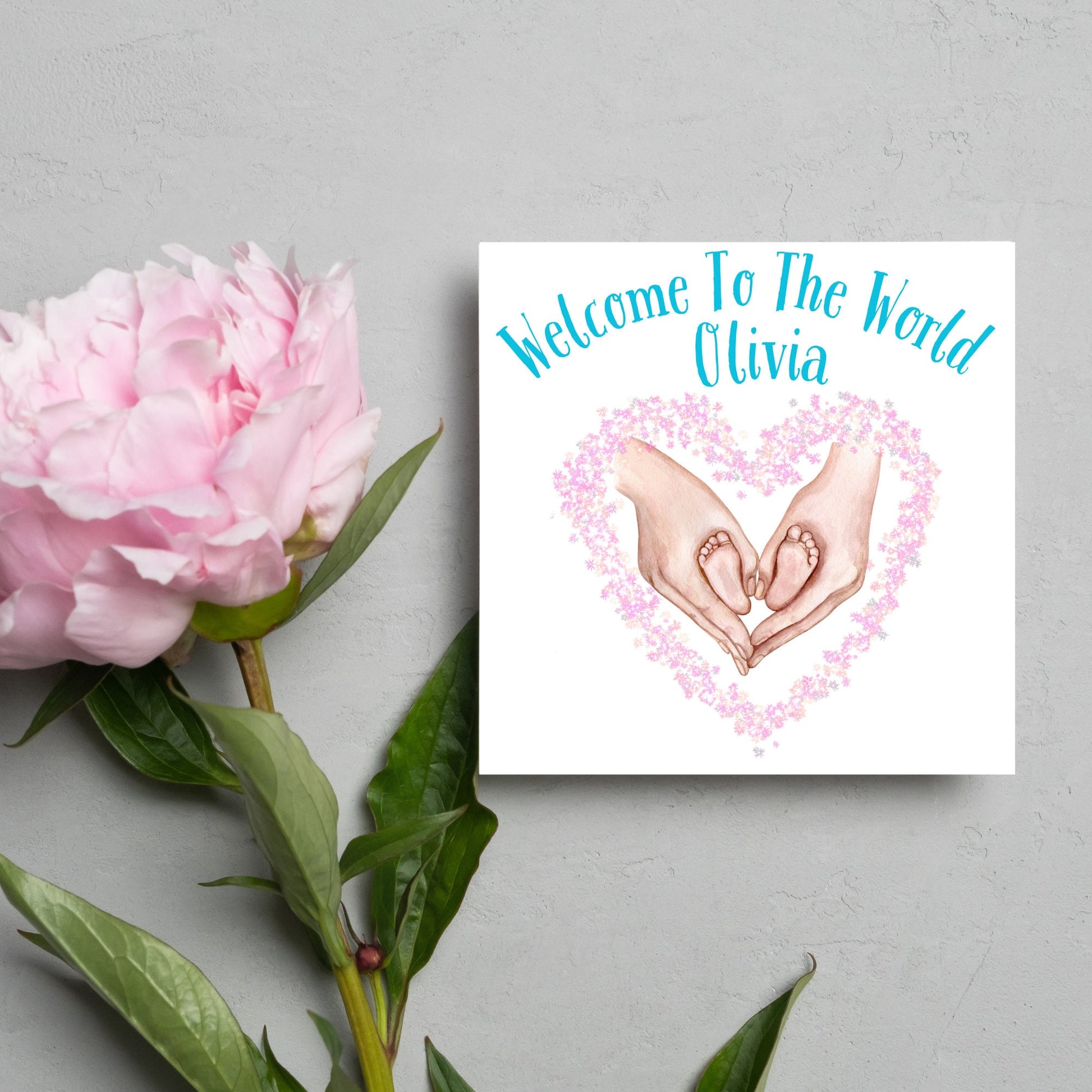 image of a square card with an illustration of hands holding newborn feet in a heart shape surrounded by pink flowers. Above the image is blue text that reads &#39;welcome to the world olivia&#39; in a handwritten style font