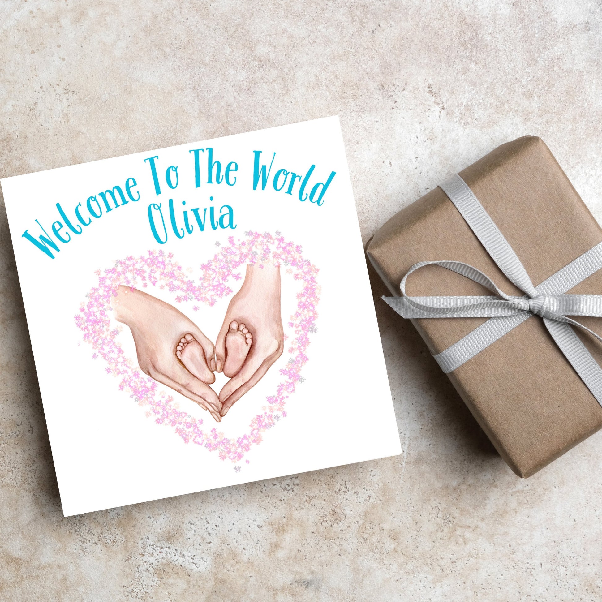 image of a square card with an illustration of hands holding newborn feet in a heart shape surrounded by pink flowers. Above the image is blue text that reads &#39;welcome to the world olivia&#39; in a handwritten style font