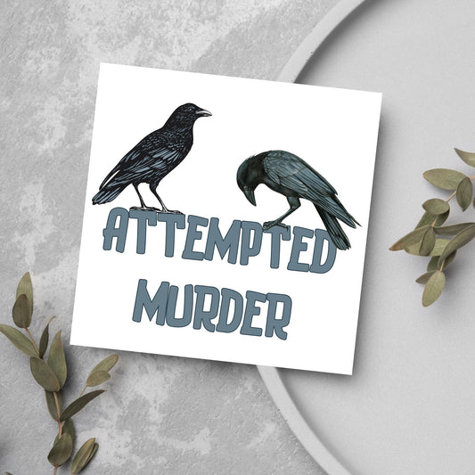 square card with the words attempted murder and 2 crows sitting on top of the text (a murder is the collective name 3 or more crows)