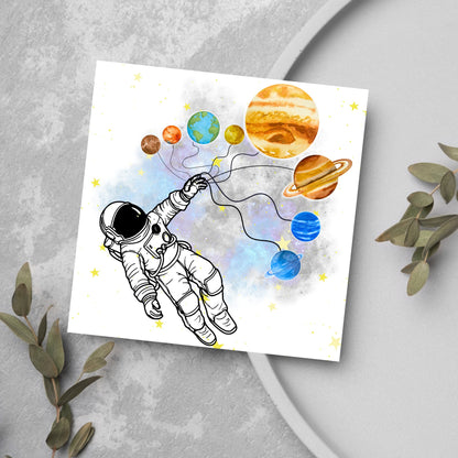 This square shaped card has an illustration of an astronauts holding the planets of the milky way in a watercolour inspired design. There is a collection of yellow stars and a watercolour nebula in the background