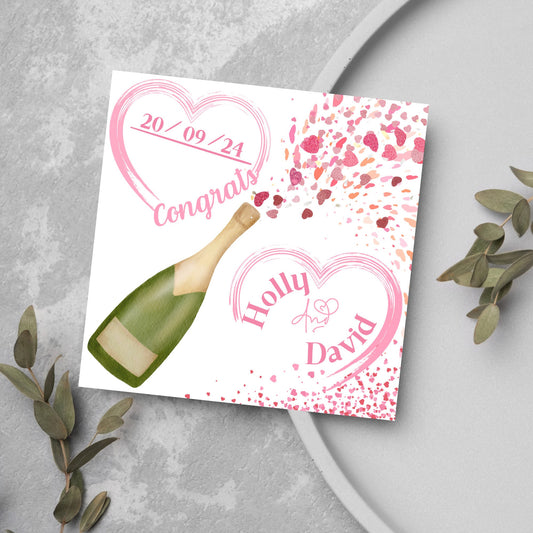 square wedding card with a bottle of champagne bursting with hearts in varying shades of pink. Above the bottle is a heart that holds the date and says contrats, and below is a repeated heart with the names of the couple inside